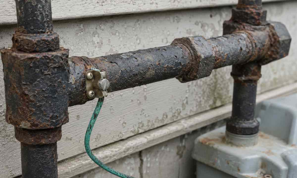 Corroded black iron gas pipe fittings with rust and pitting and oxidized copper bonding wire on an exterior wall in a coastal Delmarva setting